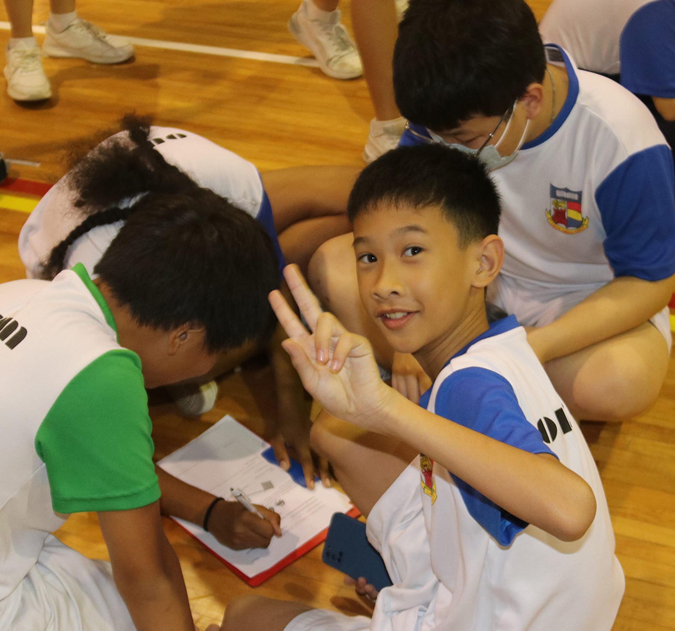 A smiling boy in a school sports uniform holds up three fingers while a teammate writes on a clipboard on a gym floor.